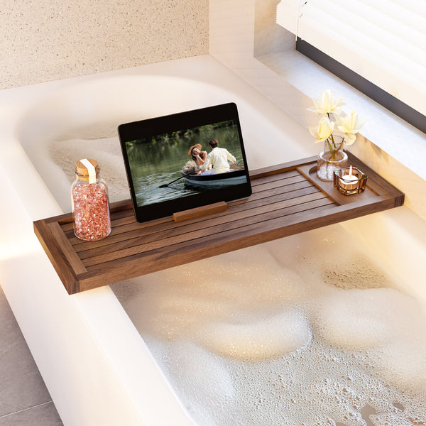 A wooden bath caddy draped over a white bathtub filled with foam, holding a tablet, a bottle of bath salts, and a small candle with flowers. No people or animals are present.