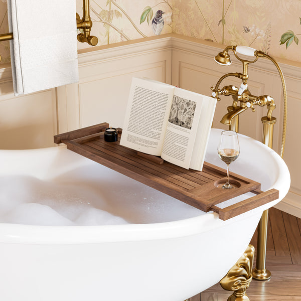 A wooden bath caddy holding an open book and a small jar rests across the edge of a white freestanding bathtub filled with bubbles. A wine glass sits in one of the caddy's cup holders, and a brass faucet and floral wallpaper are visible in the bathroom setting.