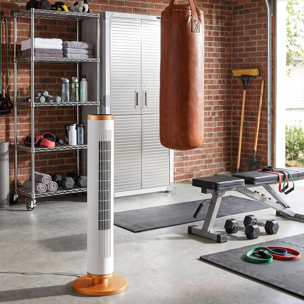 A white tower fan with copper accents stands on a concrete garage floor in a gym setting with a brick wall, punching bag, weight bench, and exercise equipment.