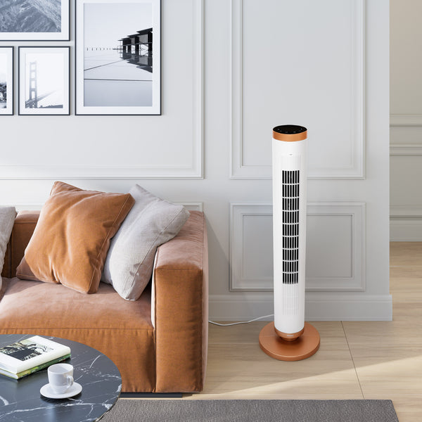 A modern white tower fan with a copper-colored top and base stands on a light wood floor in a styled living room next to a brown sofa.