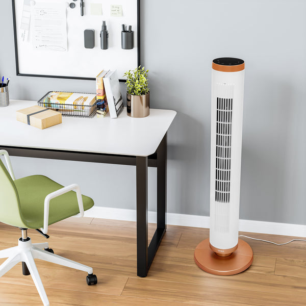 A white tower fan with a copper-colored base and top trim stands on a light wood floor next to a white desk in a modern home office setting.