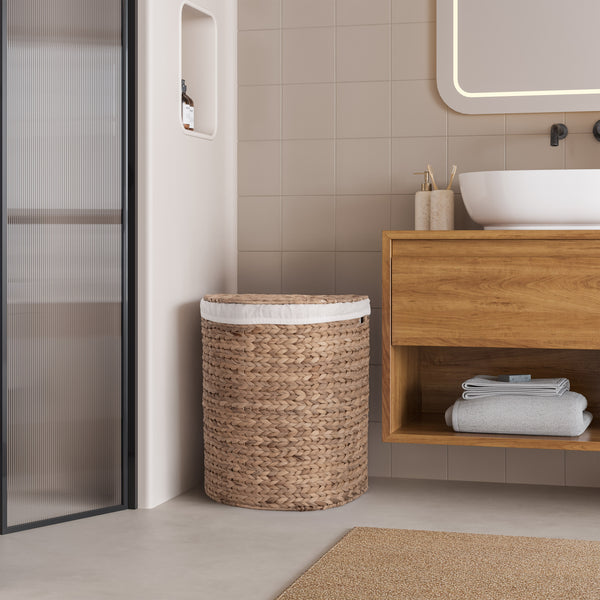 A woven natural-fiber laundry hamper with a white fabric liner, placed beside a wooden bathroom vanity with a white sink and beige tiled wall.
