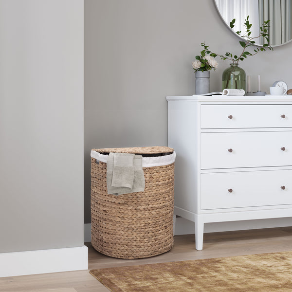 A natural woven seagrass laundry hamper with a white liner and a folded beige towel, placed next to a white dresser in a styled bedroom setting.
