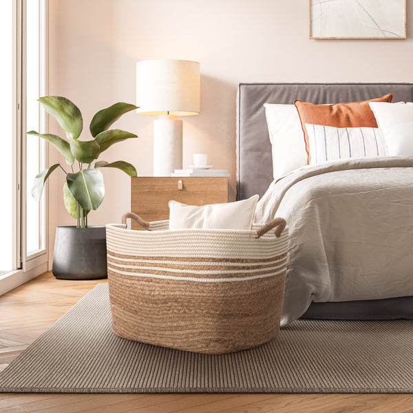 A woven water hyacinth storage basket with cream horizontal stripes sits on a striped rug in a styled bedroom setting next to a bed and a potted plant.