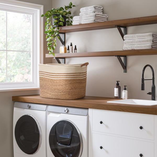 A styled laundry room corner featuring a large woven straw laundry basket on a wooden countertop, with a white washing machine and dryer, open wood shelves with folded towels, and a window in the background.
