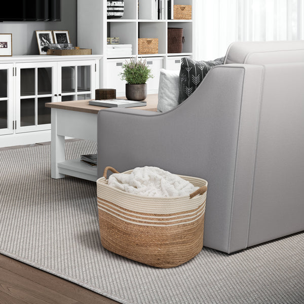 A woven water hyacinth laundry basket with cream stripes and rope handles, filled with a white blanket, placed beside a light gray sofa in a modern living room setting.