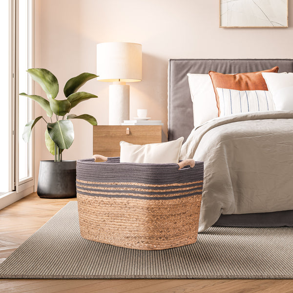 A woven storage basket with a dark gray top and natural tan woven base, placed at the foot of a bed in a styled bedroom setting.