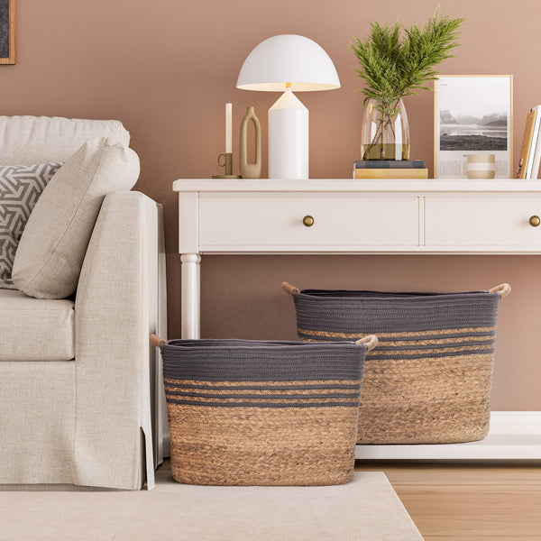 Two woven storage baskets with a dark gray rope and natural water hyacinth base, styled in a modern living room next to a sofa and console table.