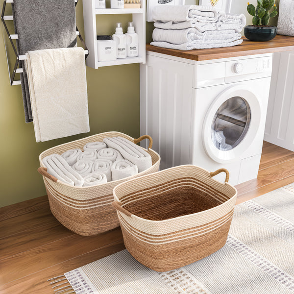 Two woven storage baskets with handles placed on a wood floor in a laundry room setting, with towels, a washing machine, and shelves in the background.