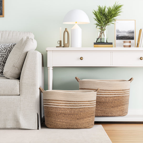 Two woven storage baskets in natural beige and brown tones with cream stripes, placed on the floor in a styled living room beside a light gray sofa and a white console table.